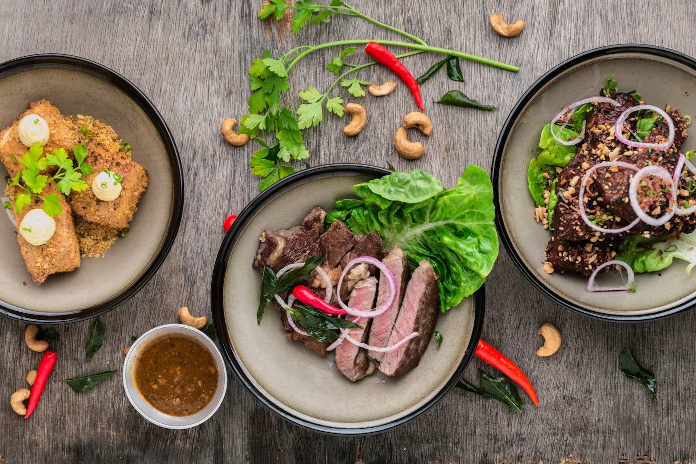 Several plated dishes garnished with fresh herbs, chillies and cashews on a dark table