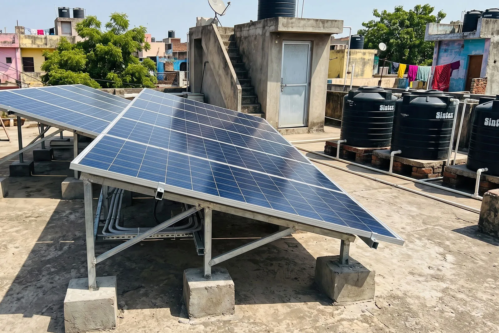 Blue solar panels on raised steel frames on the flat concrete terrace of an Indian home, with water tanks and neighbouring rooftops behind